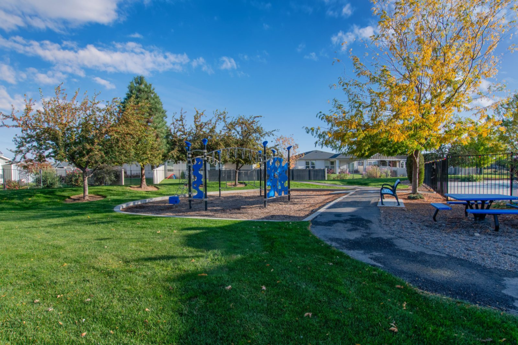 Community park with a playground, picnic tables and a paved walking path beside mature trees in the Lava Springs neighborhood in Nampa, Idaho